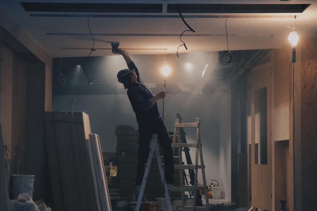 An image of someone dangerously leaning backwards on a ladder to install a lightbulb on a ceiling.